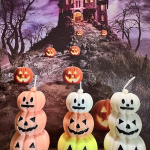 May include: Three stacked pumpkin candles, each featuring three carved jack-o'-lantern faces with black painted details. The left candle is orange, the center candle transitions from white to orange to yellow, and the right candle is white.