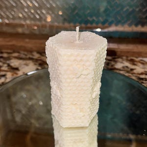 May include: A hexagonal pillar candle in an off-white color featuring a textured honeycomb pattern on its sides. The candle has a single centered wick at the top and is crafted with a wax material that displays a subtle, matte finish throughout.