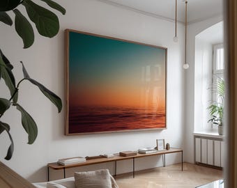 May include: A large framed photograph of a sunset over the ocean. The photograph is hanging on a white wall in a living room with a wooden floor and a white couch.