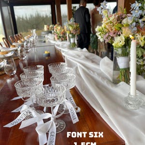 Könnte beinhalten: Eine Hochzeitstafel mit Kristall-Champagnergläsern, jeweils mit einem weißen Band mit Namen versehen. Der Tisch ist mit Blumenarrangements und einem weißen Läufer dekoriert. Der Text "FONT SIX 1,5 cm HEIGHT" ist sichtbar.