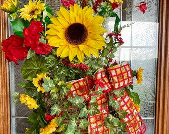 May include: A sunflower wreath with red geraniums, green leaves, and a red and white checkered ribbon with sunflowers printed on it.