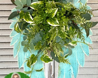 May include: A teal blue wooden angel wing wreath with a variety of green plants and greenery inside. The wreath is hanging on a wooden door.