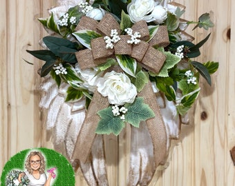 May include: A white wooden angel wing wreath with a burlap bow and white flowers. The wreath is decorated with greenery and white berries.