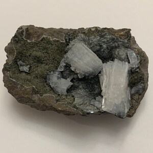 May include: A close-up of a rock with white, needle-like crystals embedded in it. The rock is a dark grey colour with green accents.