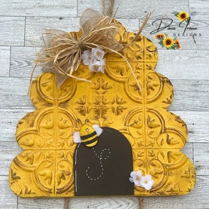 May include: A yellow and black bee-themed door hanger with a sign that reads "Bee Crossing". The door hanger is shaped like a beehive and has a textured surface. The sign is made of wood and has a distressed look.