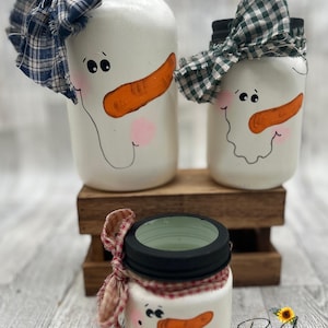 Könnte beinhalten: Drei handbemalte Schneemann-Einmachgläser mit schwarzen Deckeln. Die Schneemänner haben orange Karottennasen, schwarze Augen und rosa Wangen. Die Gläser sind mit Stoffschleifen in blauem Karomuster, grünem Karomuster und rotem Karomuster verziert. Die Gläser werden auf einem Holzständer ausgestellt.