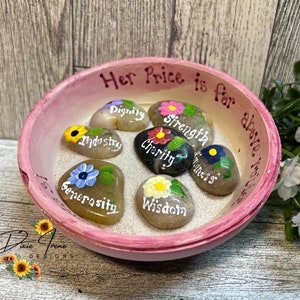 May include: A pink bowl with a white interior and the words "Her Price is far above rubies" written on the side. The bowl contains seven painted stones with words like "Dignity", "Strength", "Industry", "Charity", "Generosity", "Kindness", and "Wisdom".
