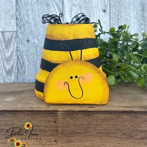 Puede incluir: Un macetero de abeja de madera pintado a mano con rayas negras y amarillas, una cara amarilla con una sonrisa y una cinta a cuadros en blanco y negro.