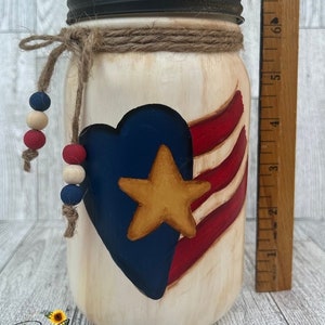 Op de afbeelding: Een witte mason jar met een zwarte deksel en een bruine touw strik. De pot is beschilderd met een rood, wit en blauw Amerikaans vlag ontwerp met een blauw hart en een gouden ster. De pot is versierd met houten en blauwe kralen.