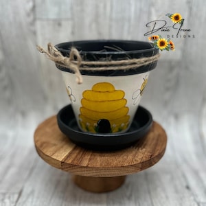 May include: A white terracotta planter with a painted beehive design and two bees. The planter has a black rim and is sitting on a wooden stand. The text "Dixie Irene Designs" is visible in the upper right corner of the image.