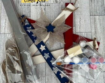 May include: DIY Craft Kit for making a patriotic star-shaped wall hanging. The kit includes wooden pieces, burlap ribbon, paint, and a paintbrush. The kit is packaged in clear plastic bags.