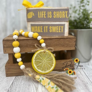 May include: A wooden sign with the text "Life is short, make it sweet" in yellow letters. The sign is decorated with a yellow lemon slice and a yellow and white beaded garland. The sign is sitting on a wooden crate.