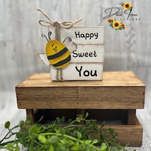 May include: Wooden block sign with the words "Happy Sweet You" painted in black. A small painted bee with black and yellow stripes is attached to the left side of the sign. The sign is sitting on a wooden crate.