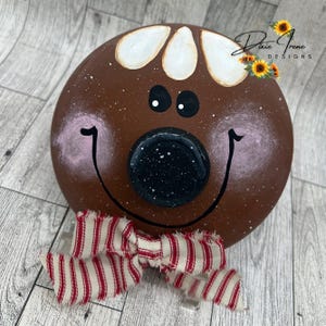 May include: A round, brown decorative piece with a painted gingerbread man face. The face has white accents, black eyes, a black nose, and a smiling mouth. A red and white striped bow is attached to the bottom. The text "Dixie Irene Designs" is visible.