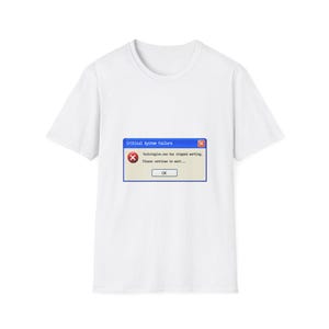 Op de afbeelding: Een wit t-shirt met korte mouwen en een blauwe computermelding in het midden. Het venster heeft de titel Critical System Failure en toont de tekst fuckstogive.exe has stopped working. Please continue to wait... met een OK-knop.