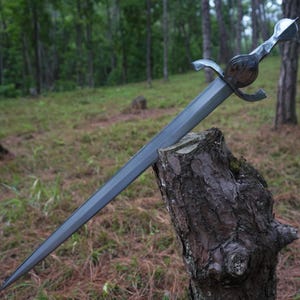 May include: A silver-toned metal sword with a double-edged blade, a central fuller, and a curved crossguard. The hilt features a rounded pommel and a textured grip, with the blade embedded into the top of a weathered, dark brown wooden tree stump.