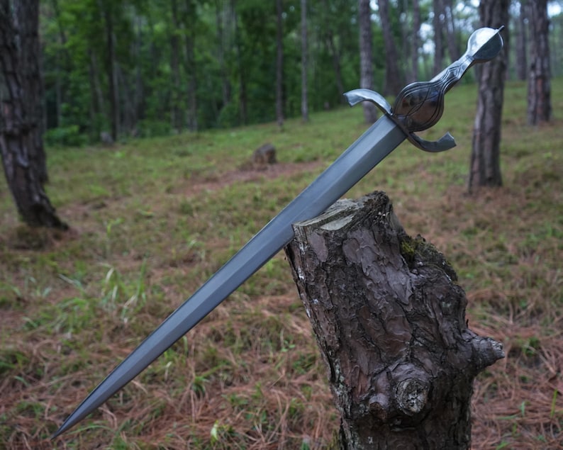 May include: A silver-toned metal sword with a double-edged blade, a central fuller, and a curved crossguard. The hilt features a rounded pommel and a textured grip, with the blade embedded into the top of a weathered, dark brown wooden tree stump.
