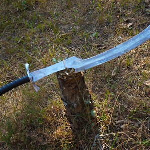Puede incluir: Una espada de color plateado con mango negro, descansando sobre un toc&oacute;n de &aacute;rbol en un &aacute;rea cubierta de hierba.