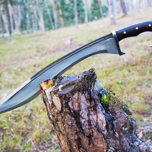 May include: A silver-colored sword with a dark wood handle and a curved blade. The sword is resting on a tree stump in a forest setting.