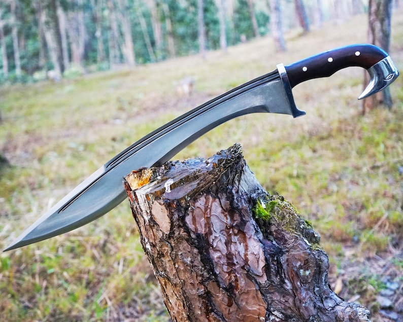 May include: A silver-colored sword with a dark wood handle and a curved blade. The sword is resting on a tree stump in a forest setting.