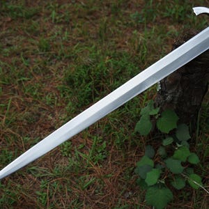May include: A long, double-edged steel sword with a pointed tip and a central ridge running down the length of the blade. The sword features a metallic crossguard and a rounded pommel at the hilt, showcasing a polished, reflective silver finish.