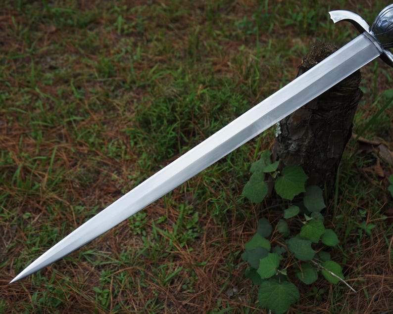 May include: A long, double-edged steel sword with a pointed tip and a central ridge running down the length of the blade. The sword features a metallic crossguard and a rounded pommel at the hilt, showcasing a polished, reflective silver finish.