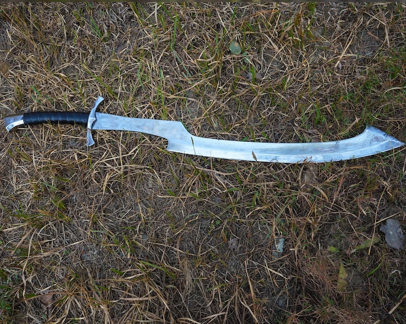 Puede incluir: Una espada plateada con mango de cuero negro yace sobre un lecho de hierba seca. La espada tiene una hoja curva y una punta afilada.