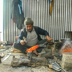 May include: A blacksmith uses a metal hammer to shape a glowing orange hot piece of steel on an anvil. The metal is heated in a nearby brick forge. The workspace is filled with various metal tools, including tongs, a kettle, and a grinder.