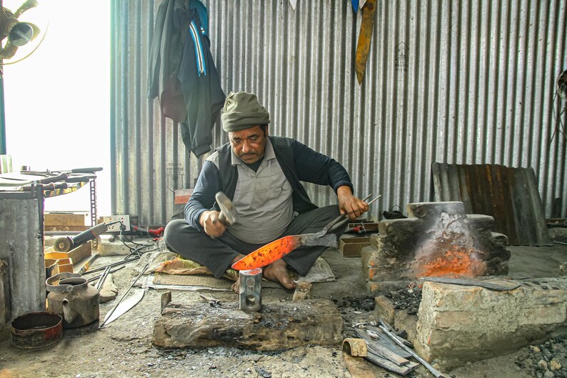 May include: A blacksmith uses a metal hammer to shape a glowing orange hot piece of steel on an anvil. The metal is heated in a nearby brick forge. The workspace is filled with various metal tools, including tongs, a kettle, and a grinder.