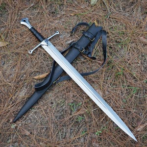 Könnte beinhalten: Ein zweischneidiges Stahlschwert mit silberfarbenem Parierstange und einem mit schwarzem Leder umwickelten Griff. Das Schwert liegt diagonal über einer schwarzen Lederscheide mit verstellbaren schwarzen Lederriemen und Metallschnallen.
