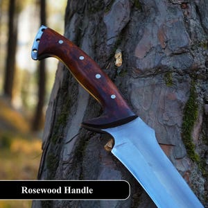 May include: A close-up of a knife with a rosewood handle and a silver blade. The knife is resting against a tree trunk. The text "Rosewood Handle" is visible at the bottom of the image.