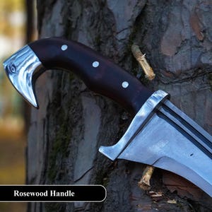 May include: A close-up of a knife with a rosewood handle. The blade is curved and has a sharp point. The handle is dark brown with white dots. The knife is resting on a tree trunk.