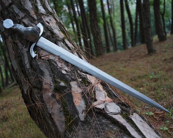 May include: A silver-colored metal sword with a double-edged blade and a curved crossguard. The handle features a rounded pommel with a metallic finish and intricate embossed detailing. The sword is positioned diagonally against a textured tree trunk.