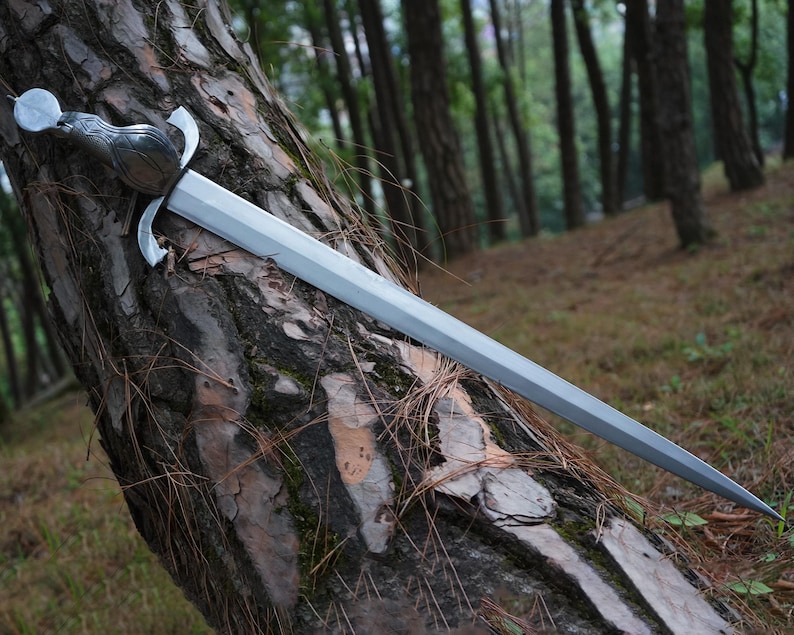 May include: A silver-colored metal sword with a double-edged blade and a curved crossguard. The handle features a rounded pommel with a metallic finish and intricate embossed detailing. The sword is positioned diagonally against a textured tree trunk.