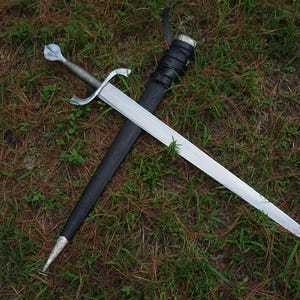 May include: A silver-toned metal sword with a pointed blade, a cross-shaped guard, and a rounded pommel. The sword is positioned diagonally across a black leather scabbard that features a silver-toned metal tip and a silver-toned metal throat.