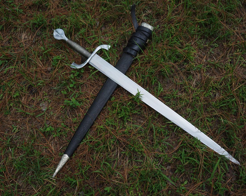 May include: A silver-toned metal sword with a pointed blade, a cross-shaped guard, and a rounded pommel. The sword is positioned diagonally across a black leather scabbard that features a silver-toned metal tip and a silver-toned metal throat.
