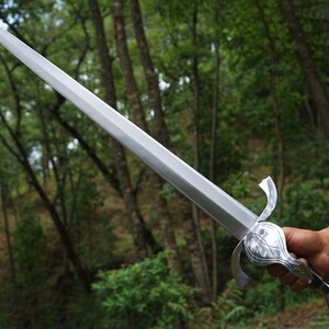 May include: A silver-colored metal sword featuring a long, straight, double-edged blade that tapers to a sharp point. The hilt includes a curved crossguard and a rounded pommel, showcasing a polished, reflective finish throughout the entire piece.