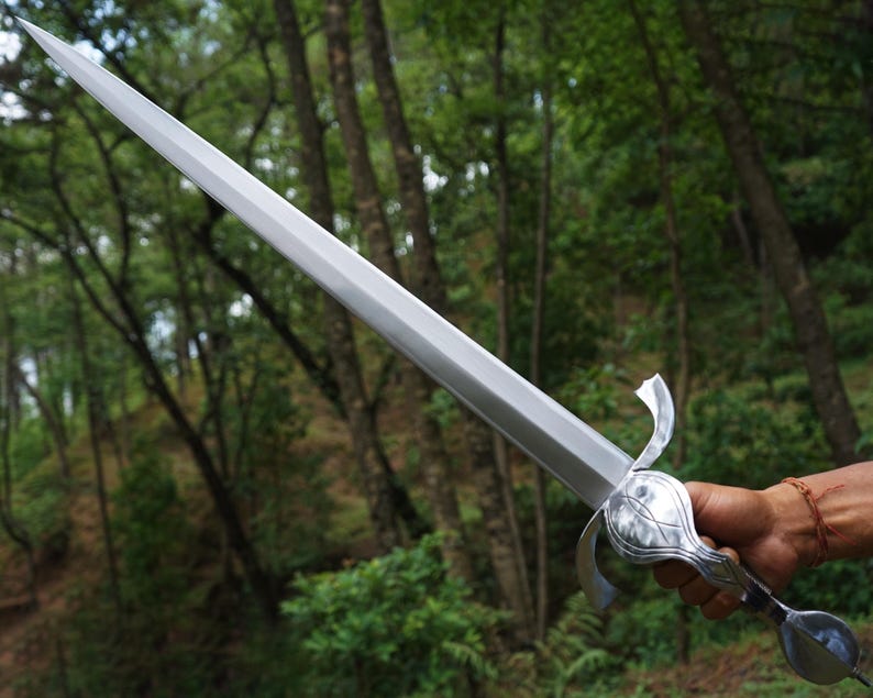 May include: A silver-colored metal sword featuring a long, straight, double-edged blade that tapers to a sharp point. The hilt includes a curved crossguard and a rounded pommel, showcasing a polished, reflective finish throughout the entire piece.