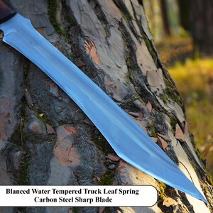 May include: A silver-colored knife with a curved blade and a wooden handle. The knife is resting on a tree trunk. The text "Blanced Water Tempered Truck Leaf Spring Carbon Steel Sharp Blade" is visible below the knife.