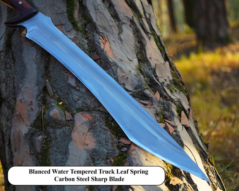 May include: A silver-colored knife with a curved blade and a wooden handle. The knife is resting on a tree trunk. The text "Blanced Water Tempered Truck Leaf Spring Carbon Steel Sharp Blade" is visible below the knife.