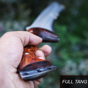 May include: A close-up of a wooden handled knife with a full tang. The knife is held in a hand with a blurred green background.