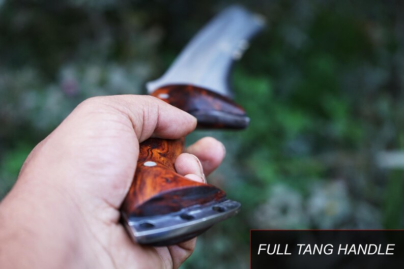 May include: A close-up of a wooden handled knife with a full tang. The knife is held in a hand with a blurred green background.