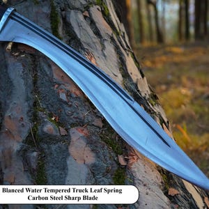 May include: A silver-colored, curved blade with a black handle. The blade is resting on a tree trunk. The text "Blanced Water Tempered Truck Leaf Spring Carbon Steel Sharp Blade" is printed below the blade.
