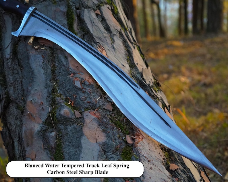 May include: A silver-colored, curved blade with a black handle. The blade is resting on a tree trunk. The text "Blanced Water Tempered Truck Leaf Spring Carbon Steel Sharp Blade" is printed below the blade.