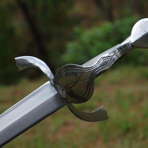 May include: A silver-toned metal sword featuring a straight, double-edged blade and a complex hilt. The hilt includes a curved crossguard, a textured grip with geometric detailing, and a rounded pommel with a small threaded metal bolt at the end.