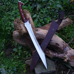 May include: A silver sword with a brown wooden handle and a leather sheath. The sword is resting on a piece of wood with green foliage in the background.