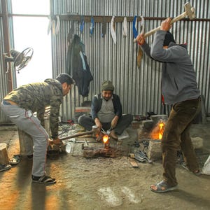 May include: Three individuals working in a metal workshop. One person stands and swings a large sledgehammer toward a glowing hot piece of metal on an anvil. Another person holds the metal with tongs, while a third person assists nearby.