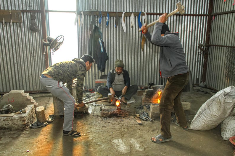 May include: Three individuals working in a metal workshop. One person stands and swings a large sledgehammer toward a glowing hot piece of metal on an anvil. Another person holds the metal with tongs, while a third person assists nearby.