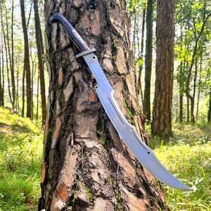 May include: A curved, silver-colored sword with a dark wooden handle and a silver guard, leaning against a tree trunk in a forest setting. The blade has a unique, curved design.