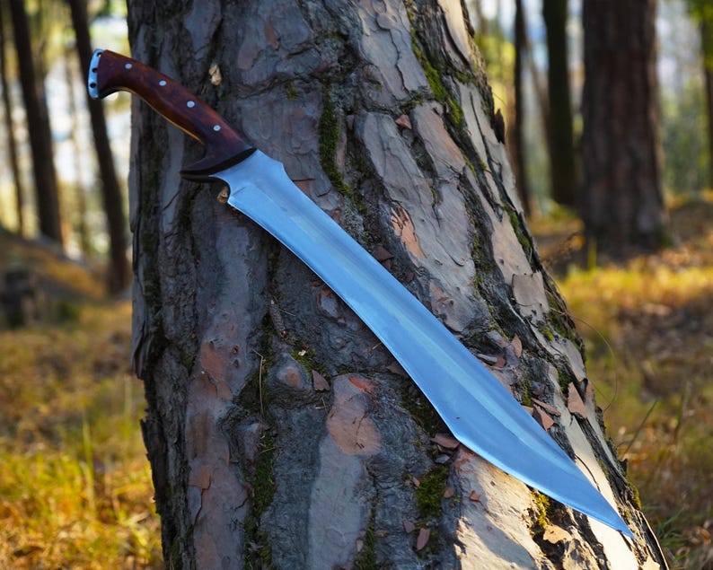 May include: A long, silver-colored sword with a wooden handle. The sword is leaning against a tree trunk in a forest setting.